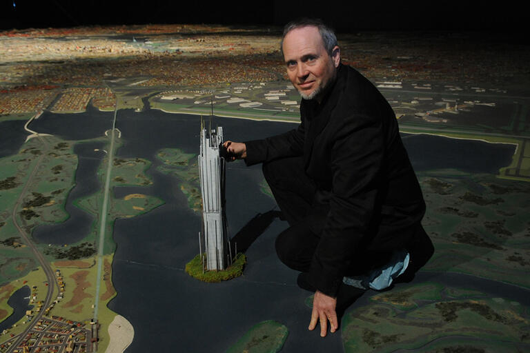 A man squats next to a detailed scale model of a cityscape, placing his hand on a tall miniature building.