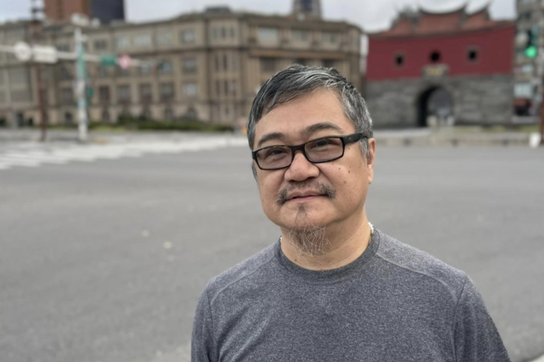 Middle-aged man with glasses standing on a street in front of an old red and gray stone gate with a large archway.