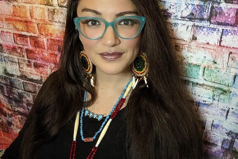 A woman with dark hair, green glasses and beaded jewelry wears a black top, looks towards the camera