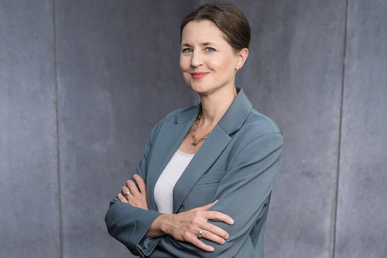 Middle-aged woman with crossed arms, wearing a teal blazer and white top, standing against a gray backdrop.