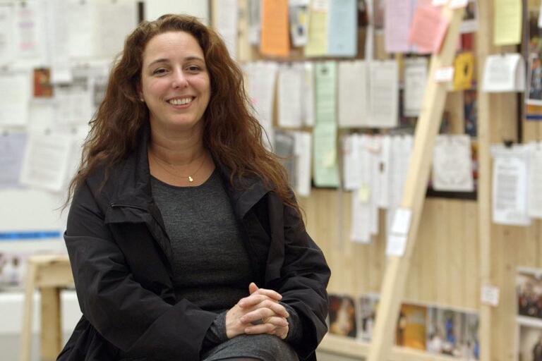 A smiling woman seated in front of a wall covered with various pinned documents and papers.