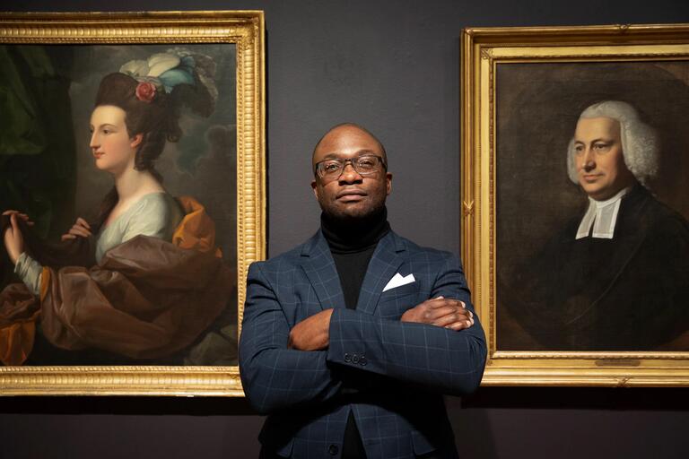 A man stands with his arms crossed between two framed portraits.