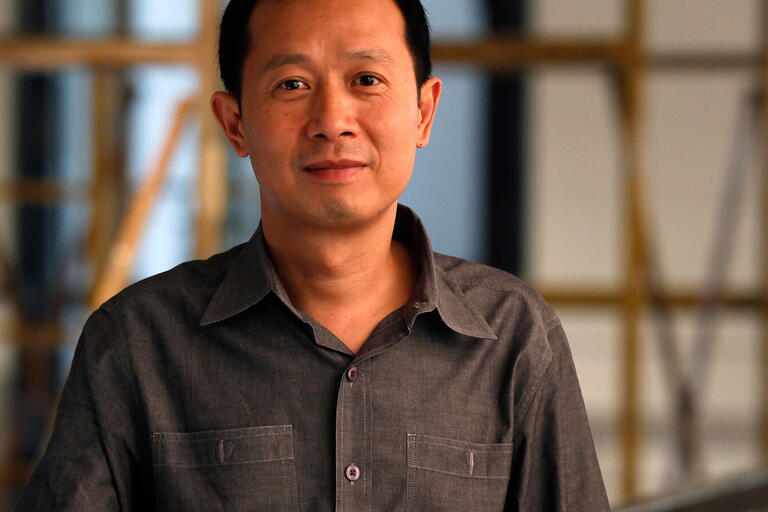 Close-up of a person in a dark grey button-up shirt standing indoors with an industrial background.