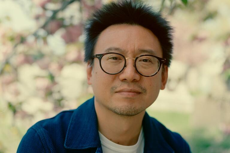 Close-up portrait of a person with dark-rimmed glasses and a blue jacket against a blurred, blooming tree background.