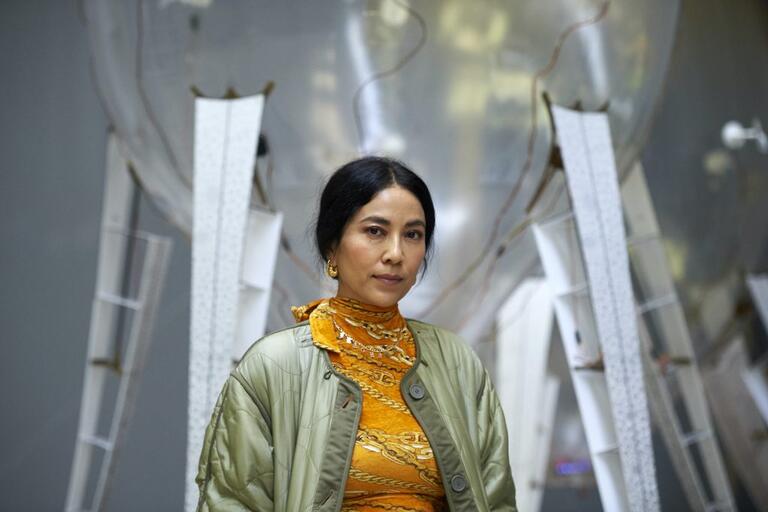 Woman in a light green jacket standing in front of a large, metallic, abstract structure.