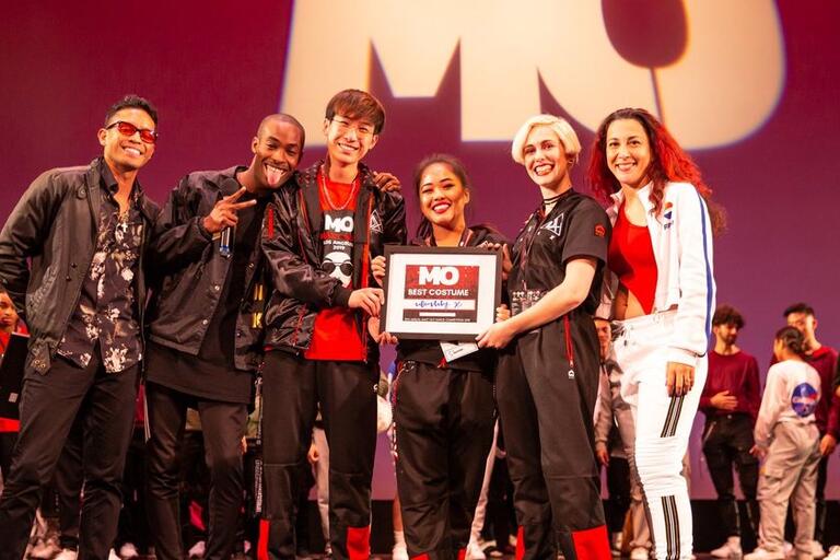 Group of six people smiling and holding a "MO Best Costume" award on stage with a crowd in the background.