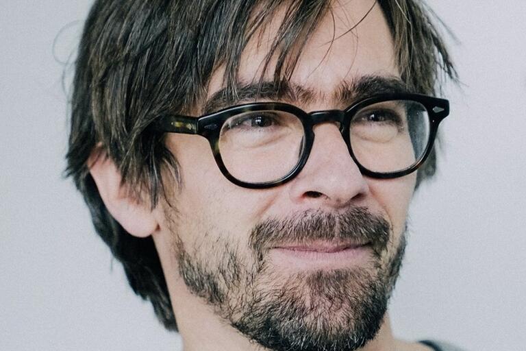 Close-up of a person with dark brown hair, beard, and tortoiseshell glasses, looking slightly to the left.