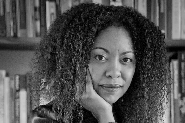 Black-and-white photo of a woman with curly hair resting her right hand on her face, with a bookshelf in the background.