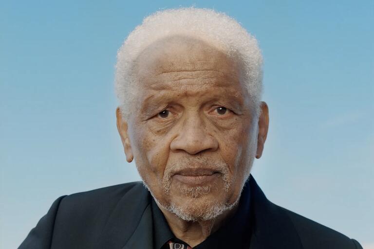 Close-up portrait of an older man with white curly hair and a beard against a blue sky background.