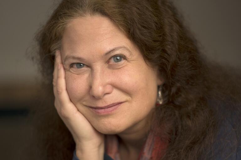 Close-up portrait of a person with long, curly, dark brown hair, smiling softly and resting their cheek on their right hand.