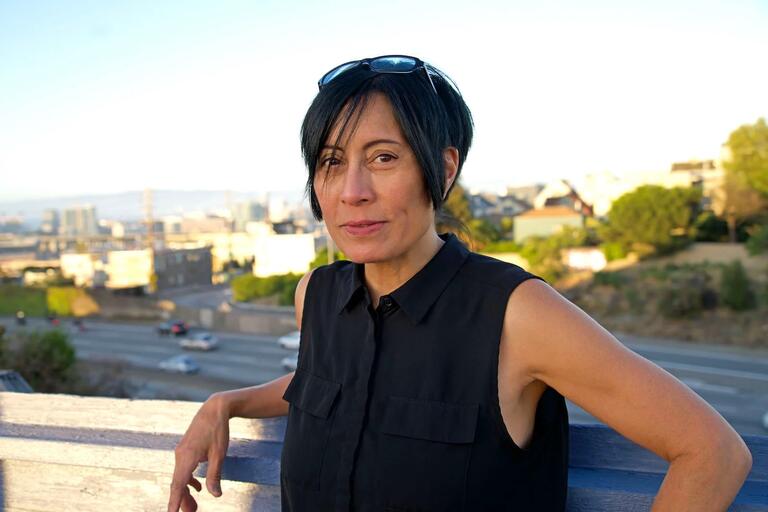 Person with short black hair, sleeveless black shirt, and sunglasses on head standing outdoors by a railing. Urban background with buildings and highway.