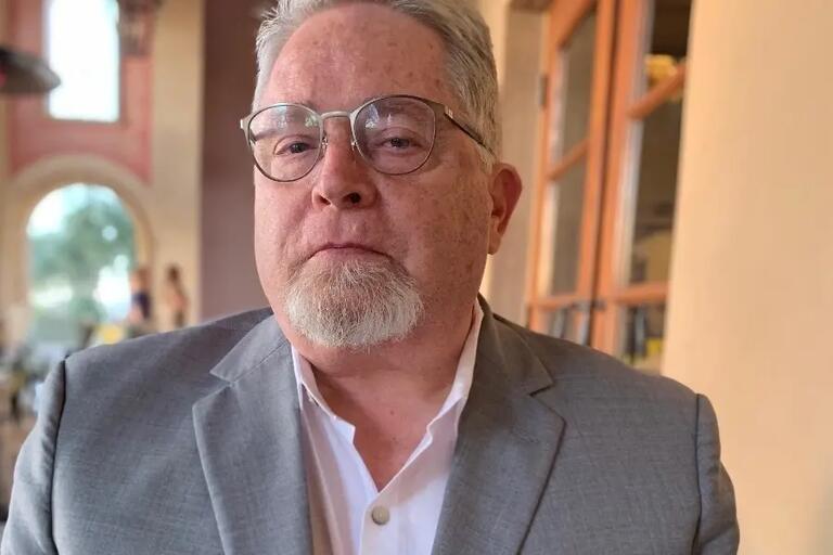Older man with glasses and a goatee in a gray suit and white shirt, standing outdoors.