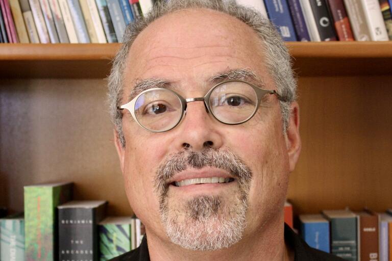 Close-up of a smiling man with gray hair and glasses in front of a bookshelf filled with books.