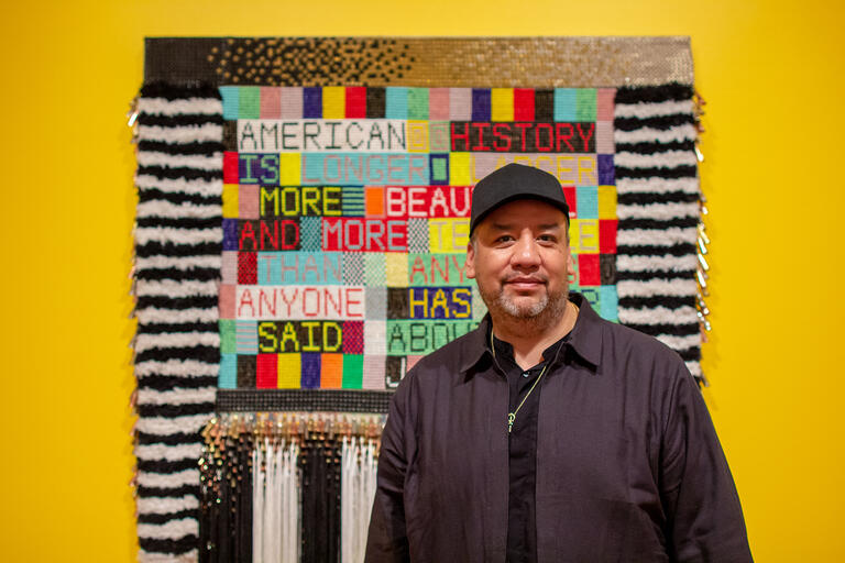 Man standing in front of a multicolored textile artwork with text on a yellow wall.