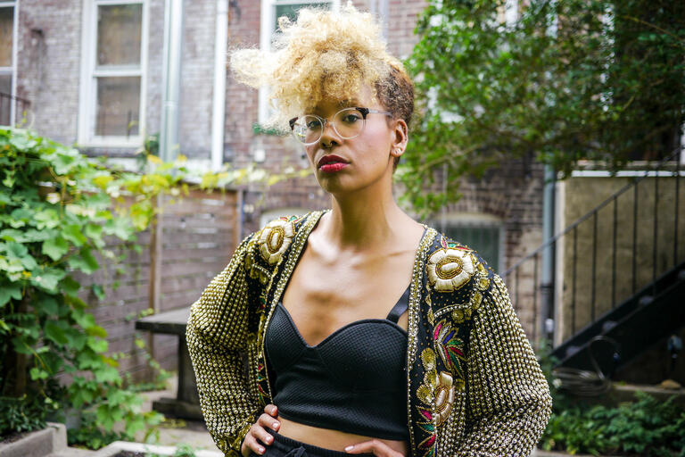 Person with curly blonde hair and glasses wearing a black top and decorative jacket standing outside with a brick building and greenery behind them.
