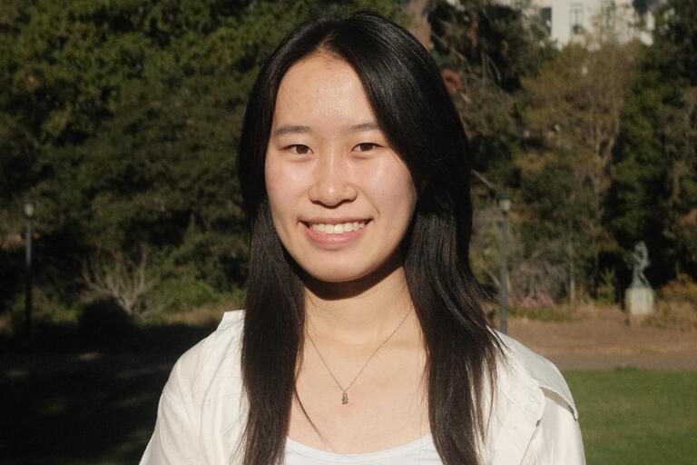 Person with long, dark hair smiling outdoors in a park with trees in the background.