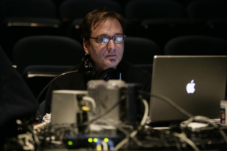Man working on a laptop in a dimly lit room with rows of empty seats in the background.