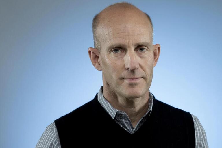 Portrait of a person in a checkered shirt and sleeveless sweater against a light blue background.