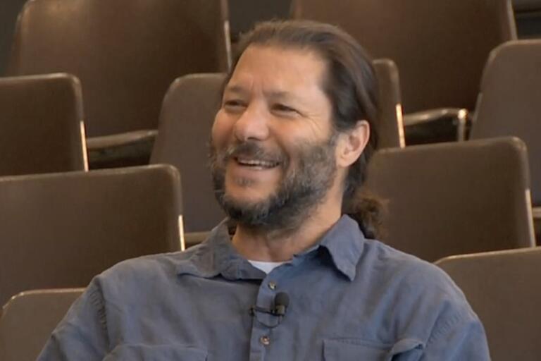 Man with a ponytail and beard, wearing a blue shirt, smiling, with empty chairs in the background.