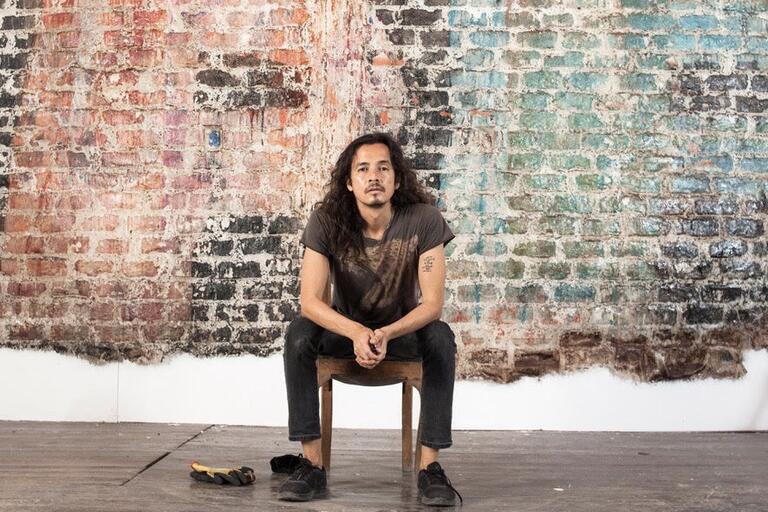 Person with long hair sitting on a wooden chair in front of a multicolored weathered brick wall.