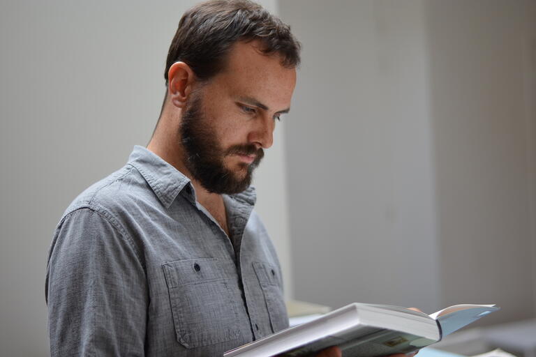 Man with short dark hair and a beard reading a book indoors, wearing a light gray shirt.
