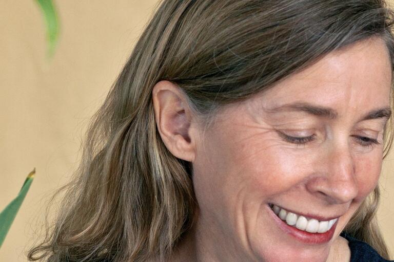 Close-up of a smiling woman looking downwards, with a beige background and a green leafy plant partially visible.