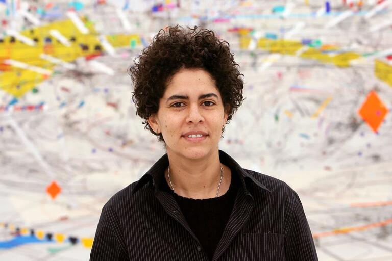 Person with short, curly hair in a black pinstriped shirt, standing in front of a colorful abstract background with geometric shapes.