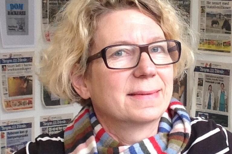 Person wearing glasses and a striped scarf in front of a wall covered with newspaper front pages.