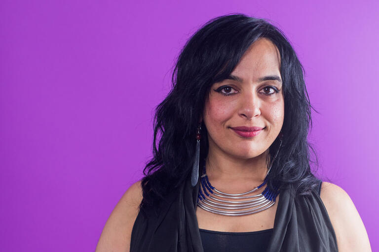 Close-up portrait of a person with black hair against a purple background, wearing a black top and a multi-layered silver necklace with blue beads.