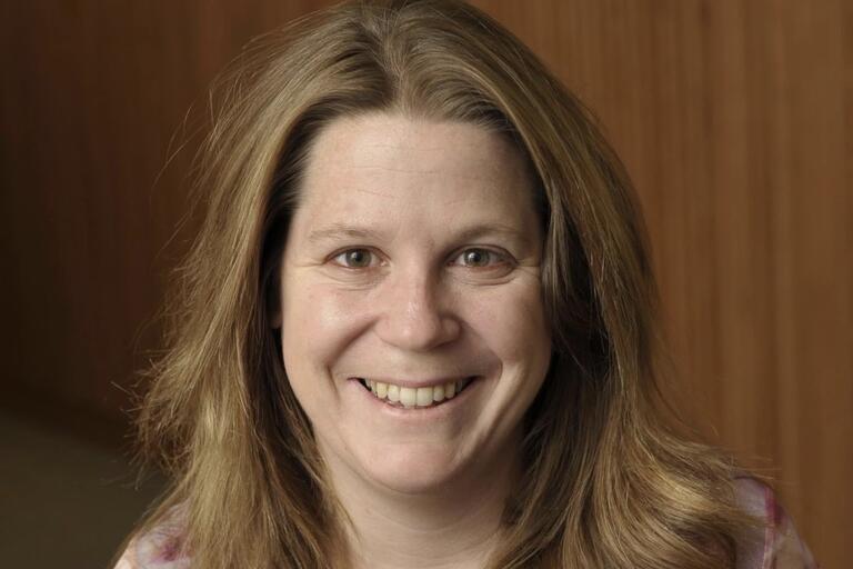 A close-up of a smiling woman with light brown hair against a wooden panel background.