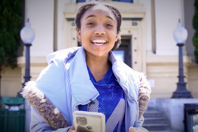 A young person smiling while holding a smartphone in front of a public building with globe-shaped street lamps.