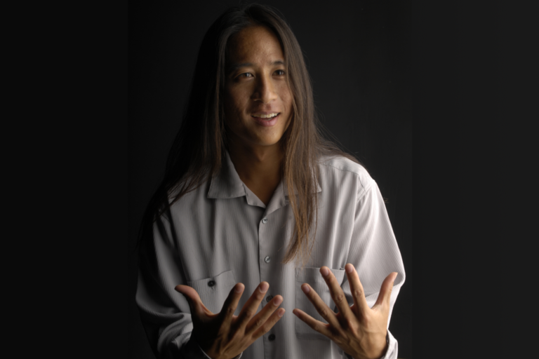 Person with long hair in a light-colored shirt, speaking with expressive hand gestures against a dark background.