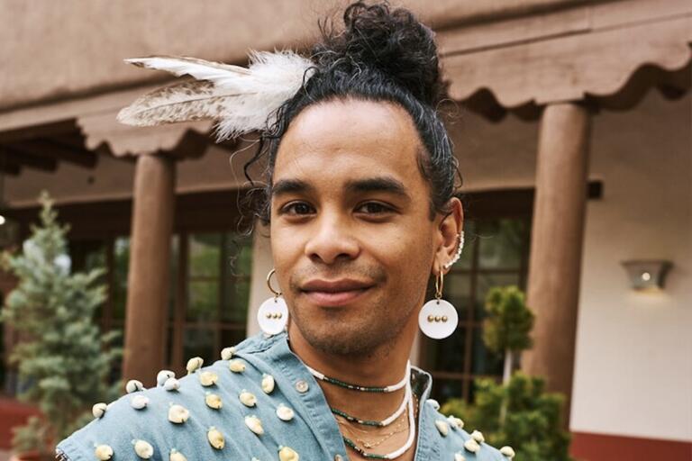 Person with curly hair in a bun, wearing a feather headdress, large circular earrings, a decorated garment, and layered necklaces, standing outdoors.