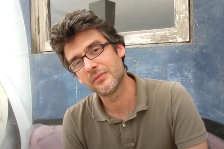 Man with glasses and short, tousled hair, wearing an olive-green polo shirt, in front of a weathered white-framed window and a blue-painted wall.