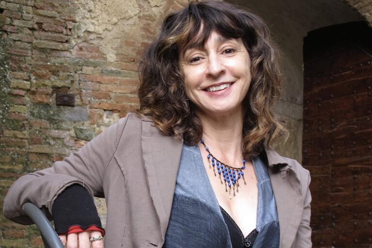Smiling woman with wavy hair wearing a light grey blazer and a blue beaded necklace, posing against a brick wall and wooden door background.