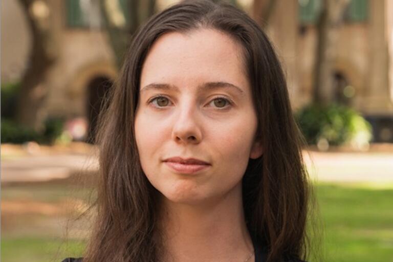 Close-up portrait of a young woman with long dark brown hair and light brown eyes standing outdoors.