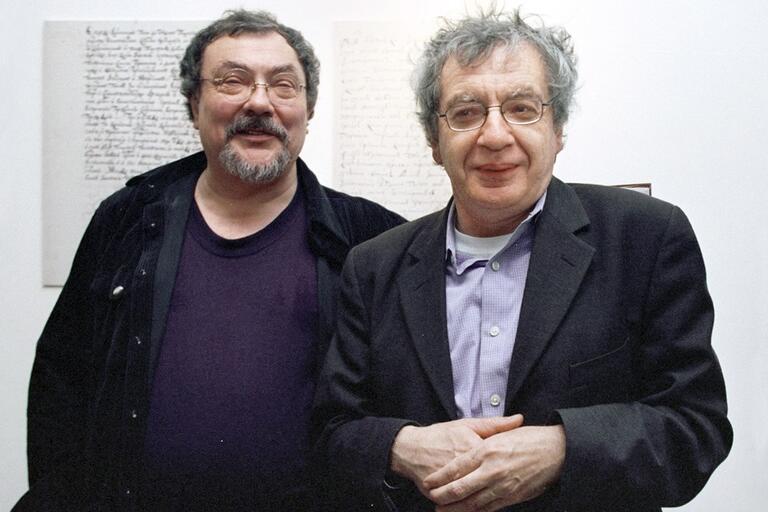 Two men standing side by side in front of handwritten text on a wall, both smiling at the camera.