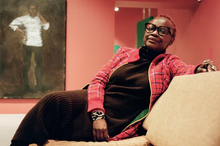 Person reclining on a light tan sofa, wearing a dark brown, ribbed sweater dress and plaid jacket, in a room with pinkish-red walls and a portrait behind them