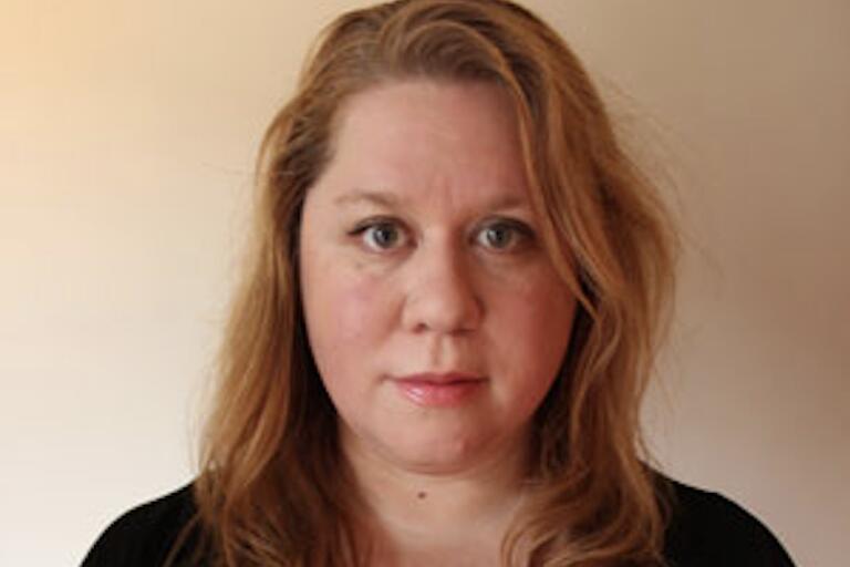 A person with long, wavy blonde hair and a black V-neck top stands against a gradient beige to tan background. They have a neutral expression and are looking directly at the camera.