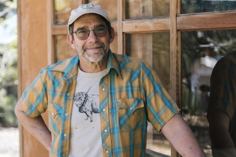 Person in a plaid shirt and cap leaning against a wooden-framed window.
