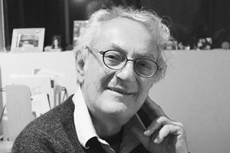 Black and white photo of an elderly person with glasses, smiling, seated in an indoor setting.