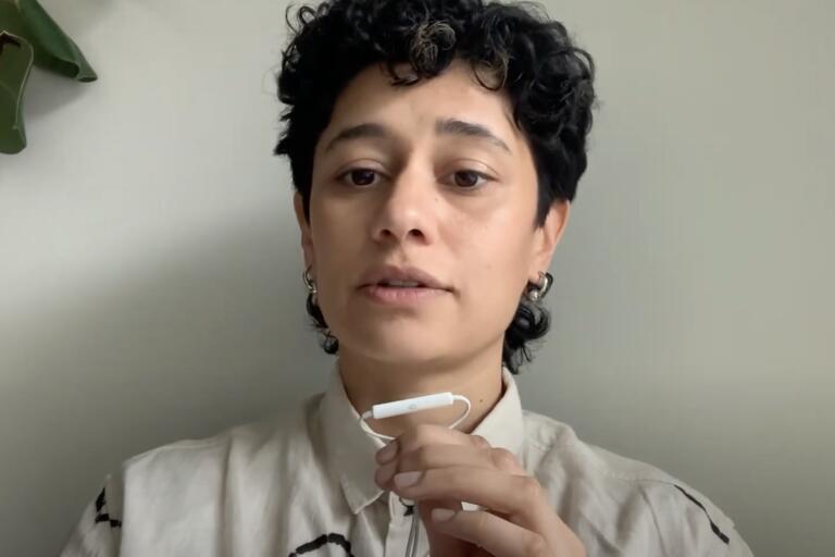 Person with curly black hair holding an earphone or small microphone near their mouth, in front of a light-colored wall and a green houseplant.