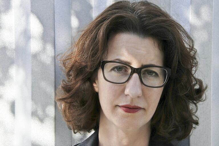 Close-up of a woman with wavy hair and black eyeglasses in front of a gray panel backdrop.