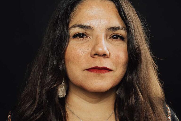 Close-up portrait of a woman with long dark brown hair, looking directly at the camera with a neutral expression.