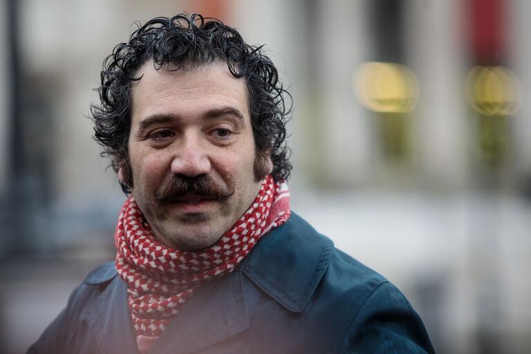 Close-up of a person with curly hair and a thick mustache, wearing a blue coat and a red and white scarf. Blurred urban background.