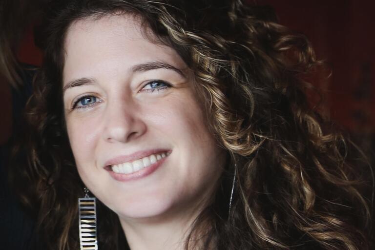 Close-up portrait of a smiling person with curly brown hair and blue eyes, wearing a long rectangular earring with horizontal stripes.