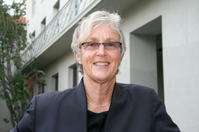 Older individual with short, light-colored hair, glasses, and a dark jacket smiling in front of a building.