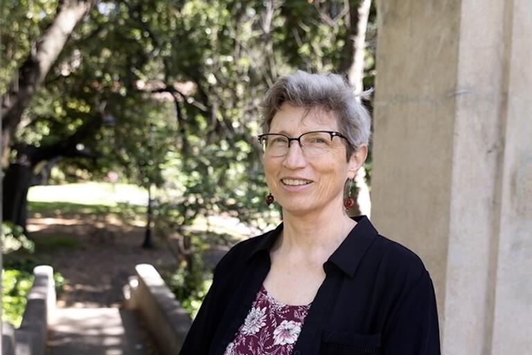Older person with short gray hair, wearing glasses and a black shirt, stands in front of a lush garden.