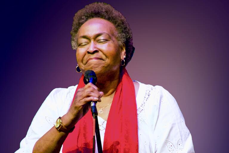 Older woman holding a microphone, wearing a white blouse and red scarf, against a purple background.