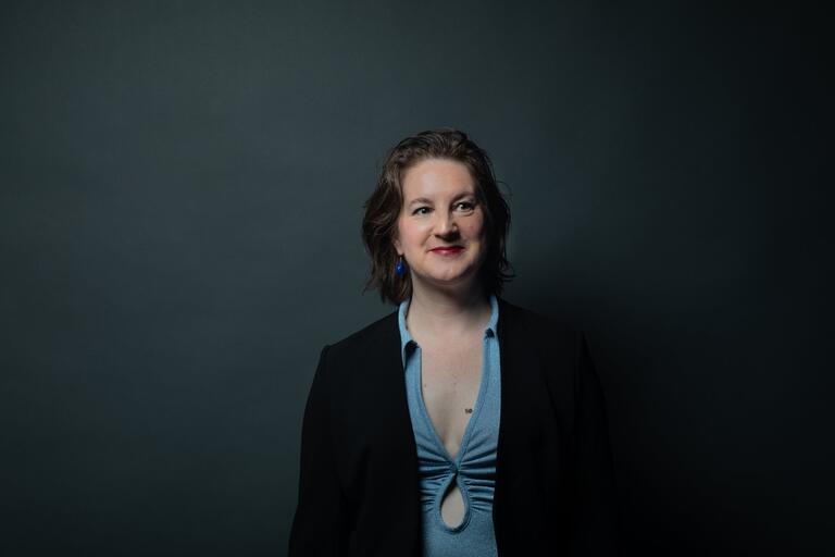 Woman with short wavy hair in a light blue top and black blazer stands against a dark background.